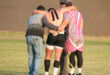 Three men support an injured rugby player with kinesiology tape on their back, walking off a grassy field. The scene conveys teamwork and care.