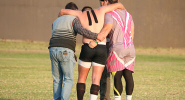 Three men support an injured rugby player with kinesiology tape on their back, walking off a grassy field. The scene conveys teamwork and care.