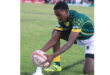 A rugby player in a green jersey and yellow socks, focused and kneeling, places a rugby ball on a kicking tee on a grassy field. It’s a calm, pre-kick moment.
