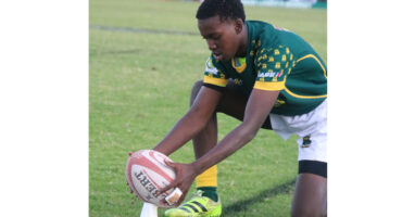A rugby player in a green jersey and yellow socks, focused and kneeling, places a rugby ball on a kicking tee on a grassy field. It’s a calm, pre-kick moment.