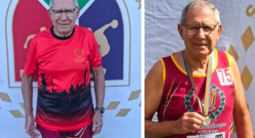 Elderly man in athletic gear stands proudly before sports-themed backdrop. He wears medals, exuding accomplishment and joy, symbolizing athletic success.