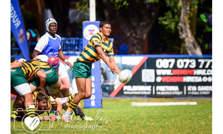 Rugby player in green and yellow stripes passes the ball mid-air during a match. Teammates are in a scrum; opponents in white nearby. Action-packed scene.