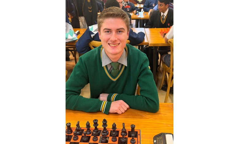 A young person in a green school uniform smiles while sitting at a chessboard in a busy classroom. The atmosphere is lively and focused.