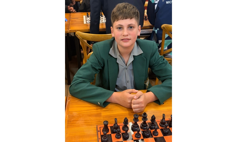 A young person in a green blazer sits at a wooden table with a chessboard. They look at the camera with a focused expression.