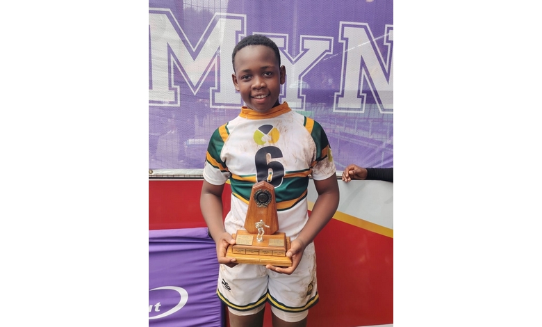 A smiling young rugby player in a green and yellow jersey, numbered 6, holds a trophy. The backdrop features large purple letters.