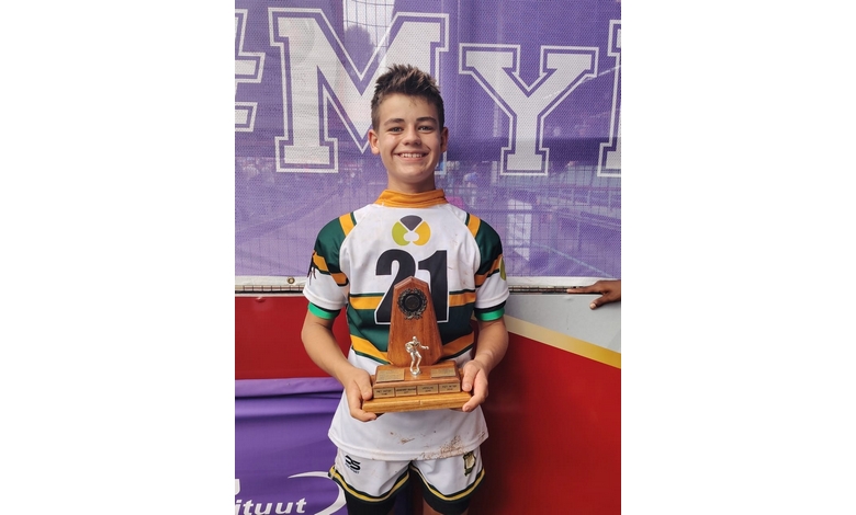 A smiling young rugby player in a white, green, and yellow jersey holds a wooden trophy. A purple banner with bold white text is in the background.