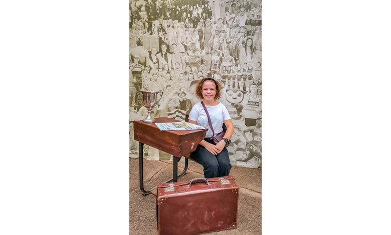 A person sits at a vintage wooden desk with a trophy, magazines, and a suitcase nearby. A sepia-toned collage of historical images forms the backdrop.