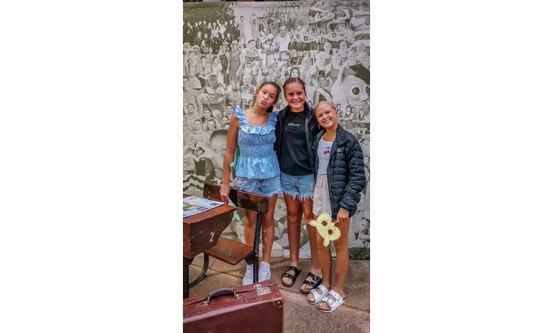 Three smiling individuals stand together in front of a mural. They're casually dressed; two wear shorts, one a dress. The scene feels joyful and relaxed.