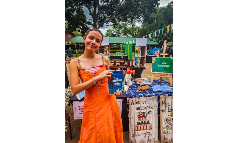 A smiling woman in an orange dress holds a book titled "Merchant of Venice" at an outdoor market stall, with colorful decorations and signs around.