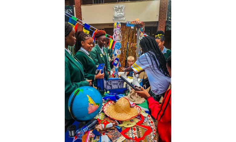 A group of students in green uniforms gathers around a colorful table with flags, a globe, and cultural artifacts. The setting is lively and educational.