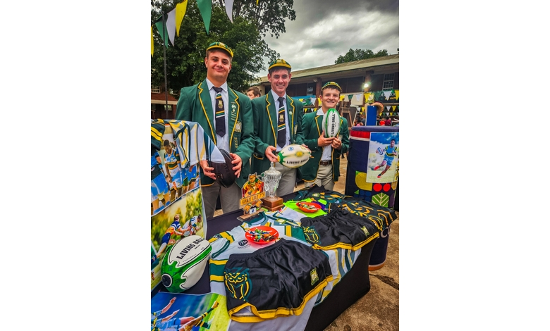Three young men in green blazers and caps stand behind a rugby-themed stall with jerseys, balls, and trophies. The mood is festive and cheerful.