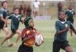 Young rugby players engage intensely in a match. A boy in a maroon and yellow jersey holds the ball, pursued by opponents in green. The scene is dynamic and focused.