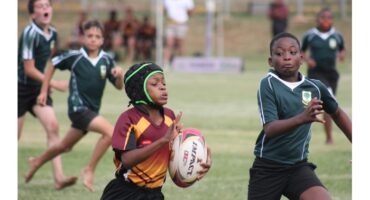 Young rugby players engage intensely in a match. A boy in a maroon and yellow jersey holds the ball, pursued by opponents in green. The scene is dynamic and focused.
