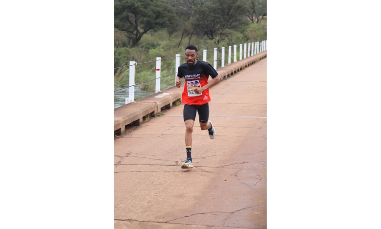 A runner in red and black athletic gear races across a concrete bridge, surrounded by lush greenery, exuding determination and focus.