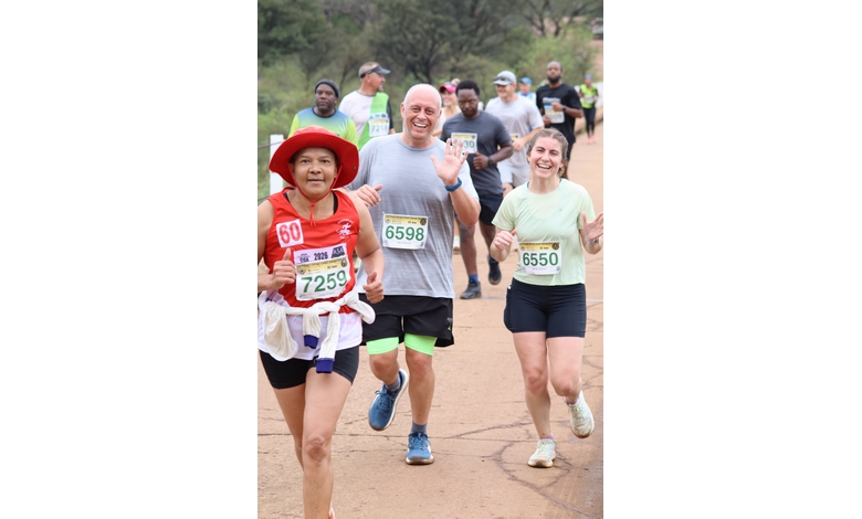 A diverse group of runners on a dirt path, smiling and mid-stride. The atmosphere is lively and energetic under a clear sky.