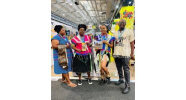 Four people in vibrant traditional attire stand smiling in a large, well-lit exhibition space. The mood is celebratory and colorful.
