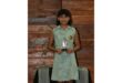 A young girl stands proudly holding a trophy, wearing a green checkered dress with a school emblem. She is in front of a wooden wall, exuding joy and achievement.