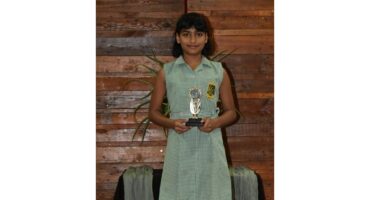 A young girl stands proudly holding a trophy, wearing a green checkered dress with a school emblem. She is in front of a wooden wall, exuding joy and achievement.
