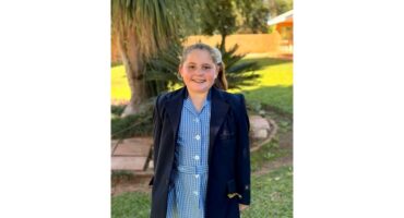 Smiling young girl in a blue checkered school dress and dark blazer stands on a garden path. Lush greenery surrounds her, evoking a cheerful mood.