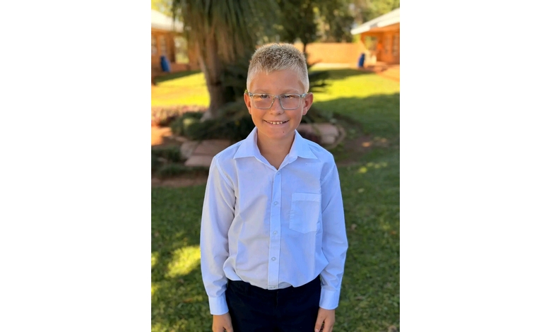 Young boy smiling, wearing glasses and a light blue button-up shirt, stands outdoors on green grass with trees and buildings in the sunny background.