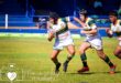 A rugby player in green and yellow stripes runs with the ball on a sunny field, pursued by two teammates, conveying action and teamwork.