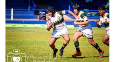 A rugby player in green and yellow stripes runs with the ball on a sunny field, pursued by two teammates, conveying action and teamwork.