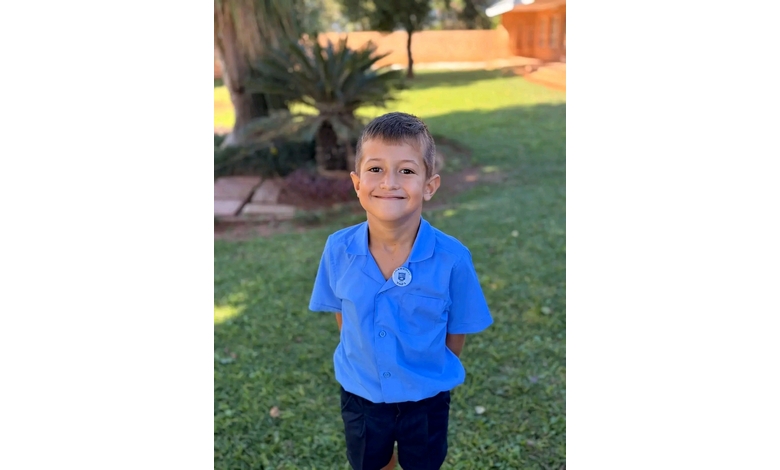 Young boy in a blue school uniform smiles widely, standing outside on green grass with trees and a wooden fence in the background. Bright, cheerful mood.