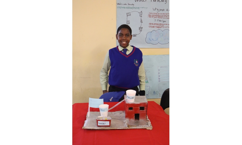 A student in a blue vest stands smiling behind a project model on a table. The model includes labeled cups and a small house structure, with educational posters in the background.