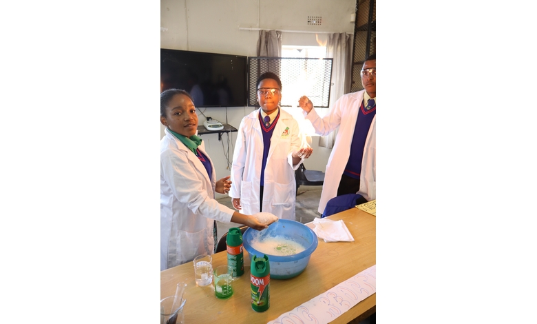 Three students in lab coats and goggles conduct a science experiment at a table. They're gathered around a blue bowl with detergent bottles nearby.