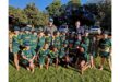 A group of smiling young children in green sports uniforms pose together on a sunny field, with two adults standing behind them. Trees and parked cars are in the background.