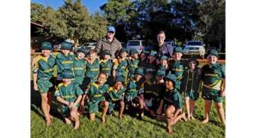 A group of smiling young children in green sports uniforms pose together on a sunny field, with two adults standing behind them. Trees and parked cars are in the background.