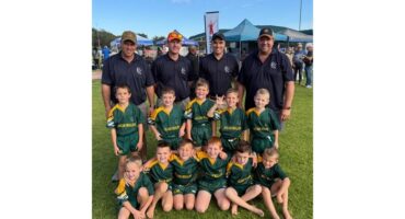 A group of young boys in green and yellow rugby uniforms pose with four adult coaches in blue shirts on a grassy field, smiling cheerfully at an outdoor event.