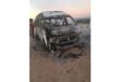 A burnt-out van on a dirt road at dusk, showing significant fire damage. A person stands nearby, looking at the vehicle with concern.