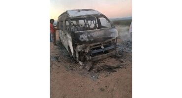 A burnt-out van on a dirt road at dusk, showing significant fire damage. A person stands nearby, looking at the vehicle with concern.