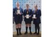 Three students in dark blue uniforms stand proudly with certificates and medals on a stage. They smile against a backdrop with a BCVO logo.
