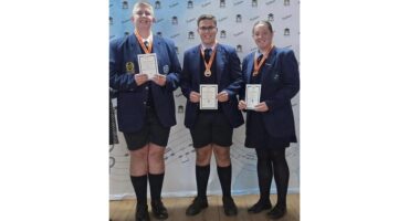 Three students in dark blue uniforms stand proudly with certificates and medals on a stage. They smile against a backdrop with a BCVO logo.
