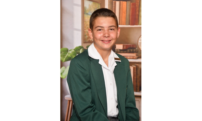 A young person in a green blazer and white shirt smiles confidently. The background features a bookshelf and plants, conveying an academic setting.