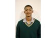 A young person stands against a plain background, wearing a green school sweater with striped trim over a collared shirt, looking confidently at the camera.
