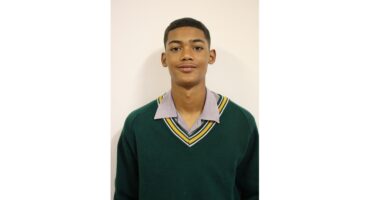 A young person stands against a plain background, wearing a green school sweater with striped trim over a collared shirt, looking confidently at the camera.