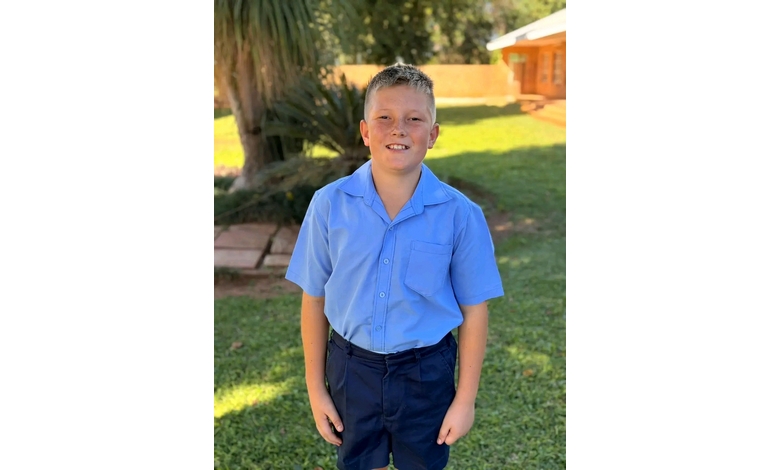 Boy in a light blue school uniform smiles outdoors, standing on green grass with trees in the background, conveying a cheerful and sunny vibe.
