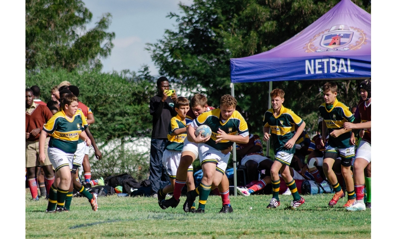 Boys in green and yellow uniforms play rugby on a grassy field, with a player in action holding the ball. A purple tent labeled "Netbal" is seen in the background.