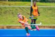 A female field hockey player in a bright orange vest lunges to hit the ball on a blue field, while a teammate watches. The scene conveys focus and action.
