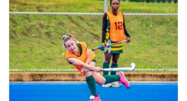 A female field hockey player in a bright orange vest lunges to hit the ball on a blue field, while a teammate watches. The scene conveys focus and action.