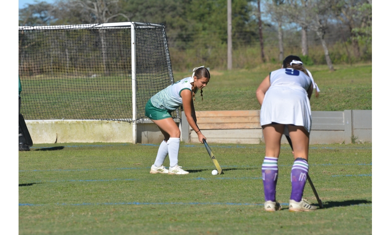 Two female field hockey players compete on a grassy field. One in green prepares to strike the ball near the goal, while the other in white defends.