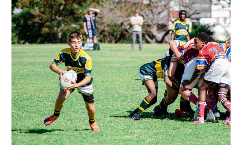 A youth rugby match with a player in green and yellow running with the ball on the grass field. Opponents are engaged in a scrum, showing teamwork.