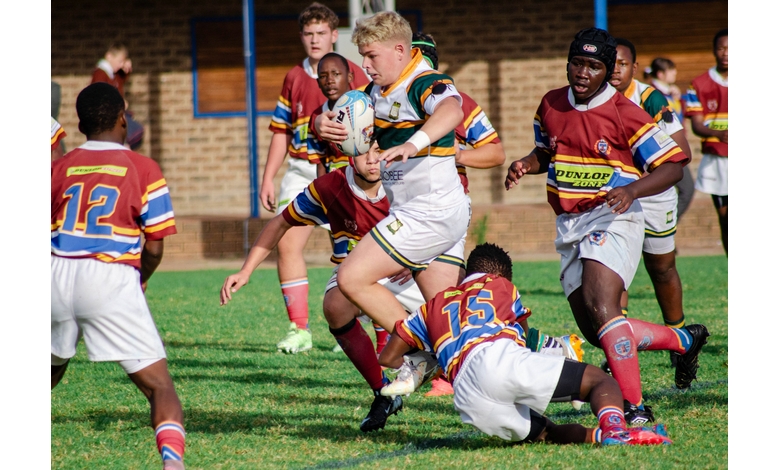 A rugby player in a white uniform runs with the ball, evading opponents in colorful jerseys during an intense school match on a sunny day.