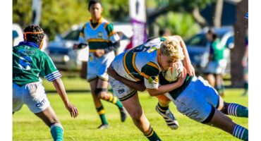 Youth rugby match with players in action. A player in a green and yellow jersey tackles an opponent with the ball, amidst others on a sunny field. Energetic scene.