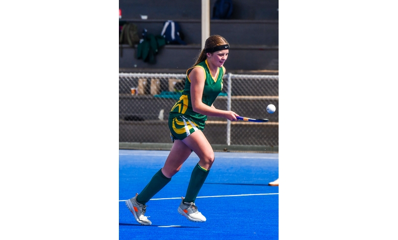 Young female athlete in green and yellow uniform plays field hockey on a bright blue turf. She focuses intently, balancing a ball on her stick.