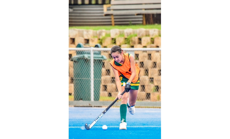Young female field hockey player in orange bib and green shorts, intensely dribbling a ball on a blue turf. Metal fence and bleachers in background.