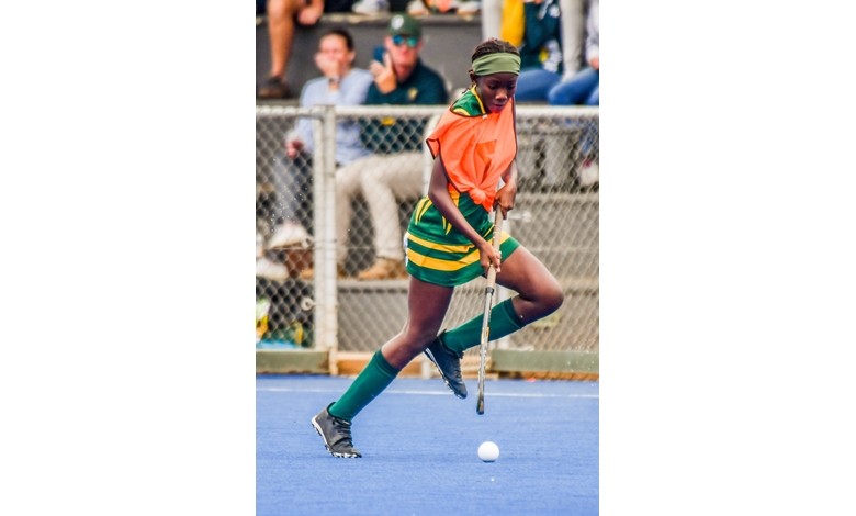 A field hockey player in green and orange moves swiftly on a blue turf, focused on the ball. Spectators and a chain-link fence are in the background.